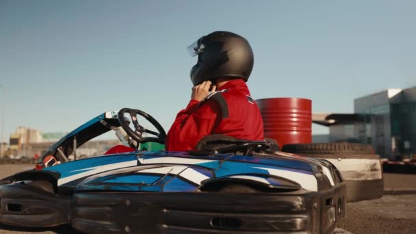 カートレースカーに座っている間 保護ヘルメットを脱ぐ制服を着たハンサムな男性ドライバー レースコースでのエクストリームゴーカートトレーニング — ストック動画