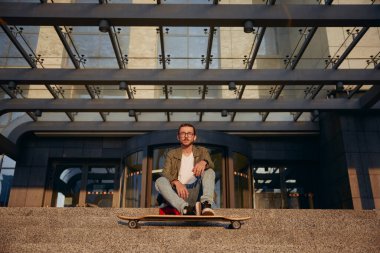 Üniversite binasının merdivenlerinde uzun tahtalı havalı bir öğrenci dinleniyor. Şehir ulaşımı için kaykay