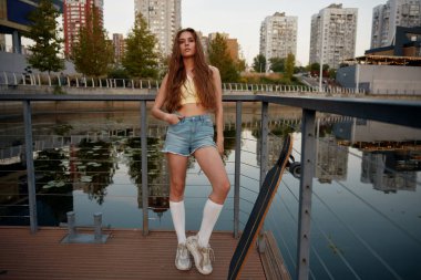 Havalı hippi kadın kaykaycı dışarıda kameraya poz veriyor. Kendine güveni tam güzel bir kadın yaz şehrinin arka planında uzun panolu günlük kıyafetler giyiyor.