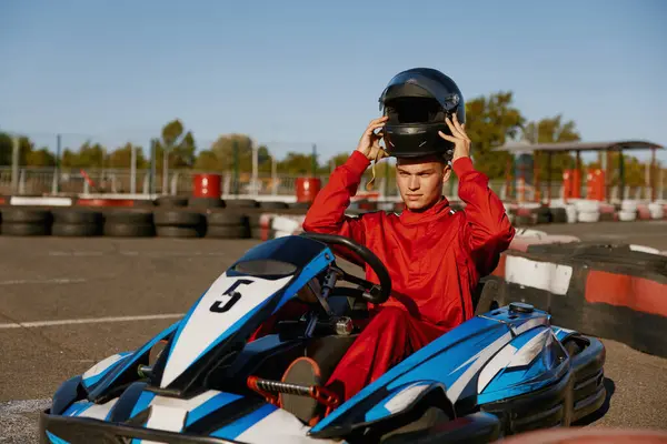 Handsome man driver wearing uniform taking off protective helmet while sitting at kart race car. Extreme go-kart training on race track
