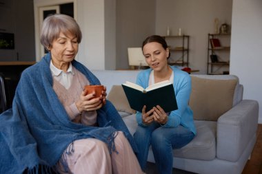 Gülümseyen yetişkin kız zihinsel bozukluğu olan yaşlı anneye kitap okuyor. Evde yaşlı ebeveynlere bakmalı ve onlara bakmalısınız.