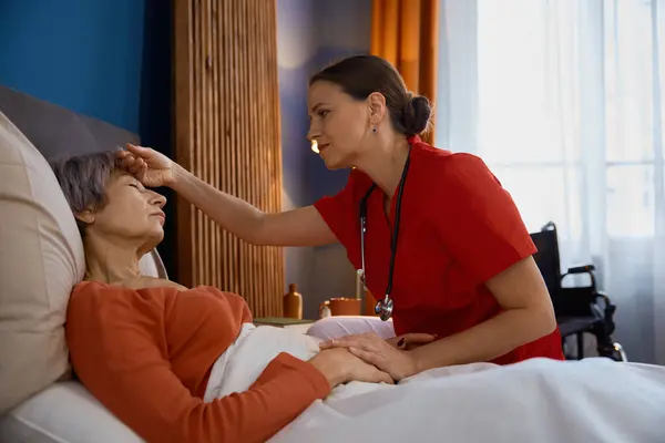女性の介護者は ベッドに横たわる老婦人の額に触れる温度を測定します 老人ホームで病気の老人を訪問する若い医師 — ストック写真