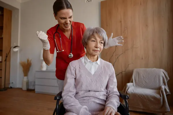車椅子の漫画のユーモラスな写真で古い女性患者のためのショットインジェクションを作るクレイジー笑顔の女性医師 — ストック写真