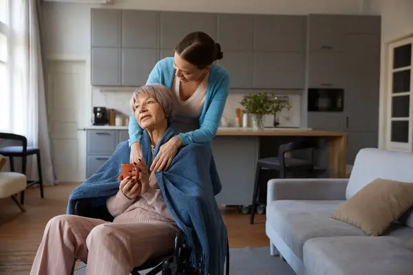 Loving adult daughter hugging old mother with disability sitting in wheelchair over cozy home interior. Happy family supporting relationship and bonding concept