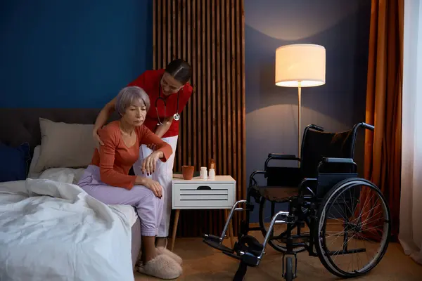 ベッドルームで悲しい不幸な老婆を助けるポジティブな家庭介護者 ベッドから車椅子に移動するために精神障害のある高齢者を支援するフレンドリーな笑顔の看護師 ストック画像
