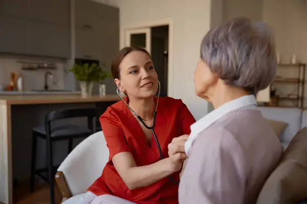 女性医師は 介護施設で高齢女性を診察しながら ステススコープを使用しています 退職後の高齢者のためのプロフェッショナルヘルスケアの身体検査 ストックフォト