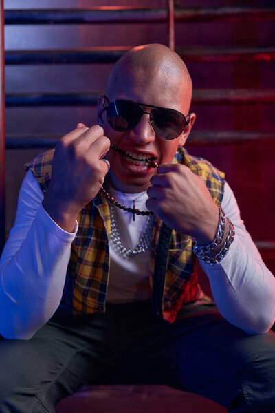 Портрет молодого красивого хипстера с лысой головой, кусающей бусы. Парень рэпера в стиле трэнди позирует перед камерой
