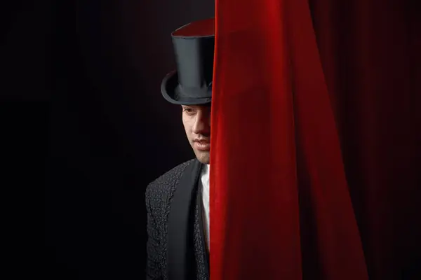 Appearing handsome man magician peeking out from behind stage drapery curtain. Young male illusionist in elegant tailcoat and top hat performing magic show and making surprise for audience