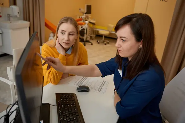 Two women doctor and patient discussing medical KT test results at clinic office. Experienced specialist explaining x-ray pointing at computer monitor