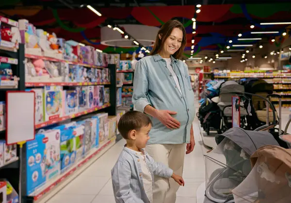 怀孕的母亲和小儿子在购物中心里选择婴儿车 为新家庭成员作准备 图库图片