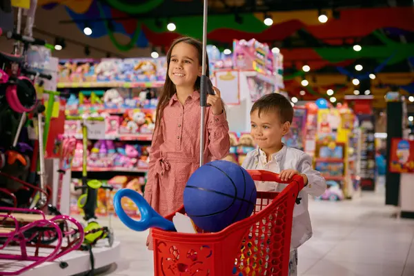 在玩具店购物的小妹妹和弟弟高兴极了 快乐的女孩和男孩手足们推着装有许多玩具的手推车的肖像 图库图片