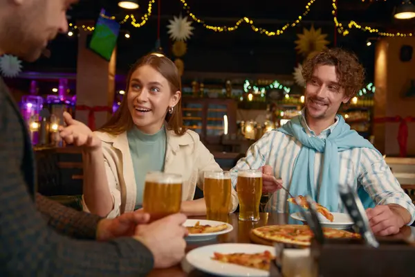 Young friends eating pizza drinking beer while rest in pub. Man and woman couple meeting old male friend sitting at table having fun conversation, sharing interesting moments of life