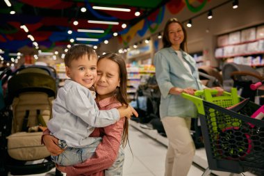 Alışveriş merkezindeki sevimli çocuklarla mutlu bir aile portresi. Sevimli kız kardeşi, sevgi ve hayranlık gösteren erkek kardeşine sarılırken hamile annesi arka planda oyuncaklarla el arabası itiyor.