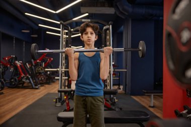 Erkek, kendinden emin okul çocuğu spor salonunda halterle vücut geliştirme antrenmanından zevk alıyor. Çocuklar için sağlıklı etkinlik