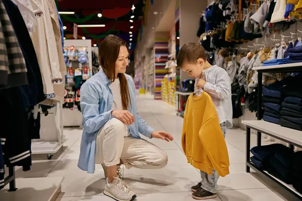 Sevgili anne ve küçük oğlu alışveriş merkezinde alışveriş yapıyor. Annem giyim mağazasındaki çocuklar için yeni moda kıyafetler seçiyor. Aile zamanı kavramı