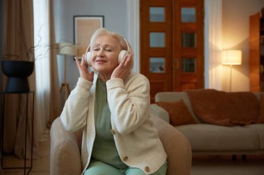 Gri saçlı, kulaklıklı, müzik dinleyen yaşlı kadın. Modern teknolojilerle emeklilik aktivitesi