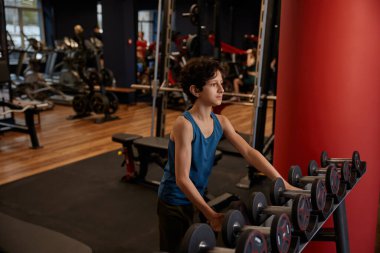 Dambıllar üzerinde dalgın bir ergen çocuk spor salonunda antrenman yapmak için ağırlık seçiminde bulunuyor.