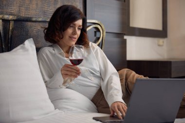 Rahatlamış iş kadını yatakta uzanıp şarap içiyor ve internette dizüstü bilgisayarla otel odasında eğleniyor.