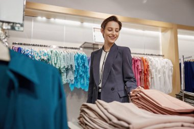 Genç zarif iş kadını modern alışveriş merkezindeki giyim mağazasında alışveriş yapıyor. Çekici bir bayan alıcı zevkle elbise vitrinleri arasında yürüyor.