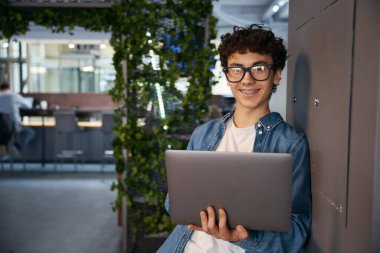 Ofiste çalışan genç bir çocuk, elinde dizüstü bilgisayarla. Gülümseyen çocuk çalışma sürecinden zevk alıyor
