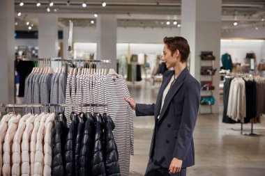 Modern alışveriş merkezindeki giyim mağazasında alışveriş yapan hoş bir iş kadını. Çekici bir bayan alıcı zevkle elbise vitrinleri arasında yürüyor.