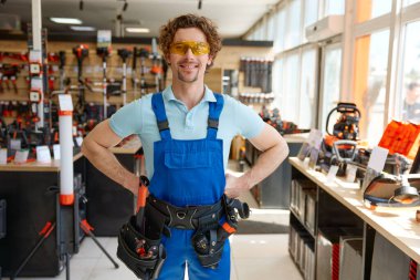 Üniformalı, koruyucu gözlüklü ve alet kemerli iyi donanımlı tamirci portresi nalbur mağazasının önünde duruyor.