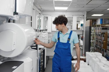 Dost canlısı gülümseyen satış danışmanı su ısıtma tankları ve kazan çeşitleriyle su deposu vitrininin başında duruyor.