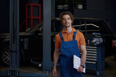 Gülümseyen araba teknisyeni elinde panoyla geziyor. Otomobil servisinde tanılama, teftiş ve tamir problemini çözmeye hazır.