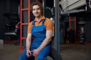 Portre mutlu gülümseyen rahat erkek tamirci garaj servisi arka planında oturuyor.