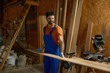 Üniformalı genç marangoz modern ekipmanlara ahşap kaplama takıyor. Deneyimli bir işçi kendi küçük mobilya fabrikasında marangozluk projesinden zevk alıyor.