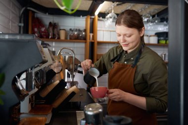 Önlüklü deneyimli kadın barista kahve bardağına taze demlenmiş içecekle süt dolduruyor. Kahve dükkanı ya da kafe personeli müşterilerine hizmet veriyor.