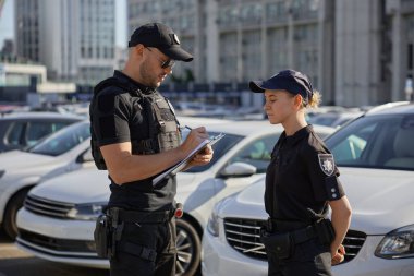 Kadın ve erkek devriye polisi park edilmiş arabalarla şehir caddesinde çalışıyor. Polis memuru panoya yazı yazıyor, genç bir polis memuruna ders veriyor.