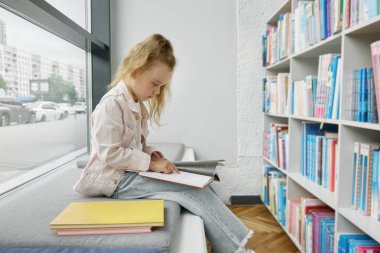 Çocuklar için halk kütüphanesinde vakit geçirirken güzel küçük kız kağıt kitap okuyor. Anaokulu öğrencisi, kitaplığın önünde oturmuş edebiyat dolu raflarla.