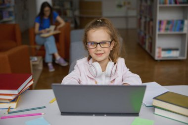 Annesi bulanık arka planda okurken, kütüphanede dizüstü bilgisayarla internet üzerinden çalışan sevimli mutlu kız çocuğu. Akıllı küçük öğrencinin portresi.