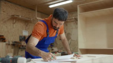 Marangoz proje taslağı ile kağıt sayfasına bakıyor ve ahşap dükkanının iç kısımlarında duran toplantı talimatlarını okuyor.