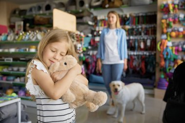 Bulanık arka planda anne ve köpekle evcil hayvan dükkanının başında duran yumuşak tavşan oyuncağına sarılan küçük kız.