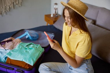 Tropik tatil köyüne gitmeden önce akıllı telefondan arkadaşlarıyla sohbet eden mutlu bir genç kadın. Güzel kız yatakta oturuyor yaz tatili için giysiler hazırlıyor.
