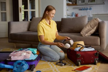 Genç gülümseyen kadın turist valiz paketliyor kulaklıkları kişisel eşyalarıyla birlikte çantaya koyuyor.