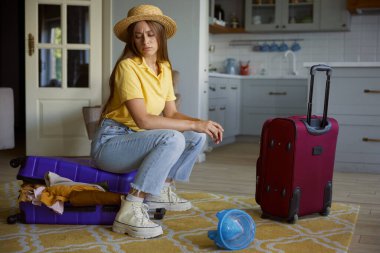 Üzgün kadın turist surat, seyahat valizine oturmuş tatil için bir sürü kıyafet hazırlamaya çalışıyor.