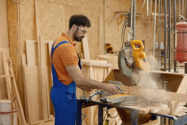 Yetenekli erkek marangoz tulum ve koruyucu gözlük takıyor. Marangozluk tezgahında yuvarlak testereyle tahta kesiyor.