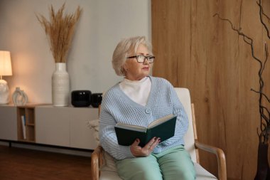 Evde sandalyede otururken kameraya poz veren gözlüklü çekici yaşlı kadın.