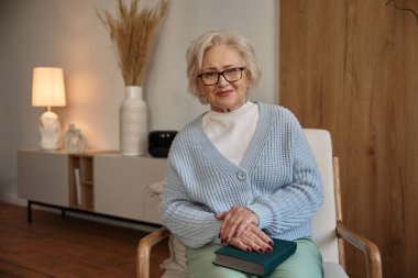 Evde sandalyede otururken kameraya poz veren gözlüklü çekici yaşlı kadın.