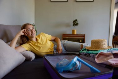 Genç bir kadın açık seyahat çantasıyla yatakta yatıyor. Tropikal tatil köyündeki lüks günleri düşleyen güzel kız.