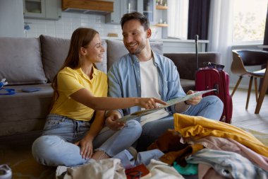 Sevgili çift, yaz tatillerinde kağıt harita okuyarak seyahat planları yapıyor. Neşeli adam ve kadın, yolculuk için hazırlanmış giysi yığınının önünde oturuyor.