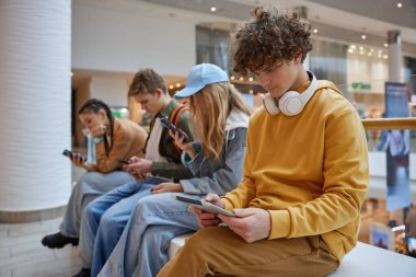 Bir grup genç, büyük şehir alışveriş merkezi ziyareti sırasında bankta otururken modern mobil cihazlar kullanıyorlar.