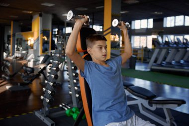 Konsantre genç çocuk spor salonunda spor yaparken dambıl kaldırıyor. Yedek kulübesinde oturan genç adam ağırlık çalışıyor kaslarını kasıyor ve güç ve güç gösteriyor.