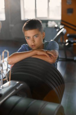 Spor kulübü halter ekipmanlarının üzerinde kameraya poz veren genç bir çocuğun portresi. Spor okulu öğrencisi halter için spor salonunun iç tasarımı üzerinde duruyor.