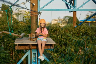 Küçük mutlu dağcı çocuk gökyüzünün altındaki şehir halat parkında maceradan tatmin oluyor. Sevimli kız kollarını açıp zevkten bağırıyor.