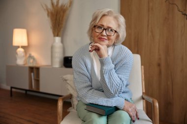 Sandalyede otururken kameraya poz veren ve gözlük takan çekici yaşlı kadın portresi.