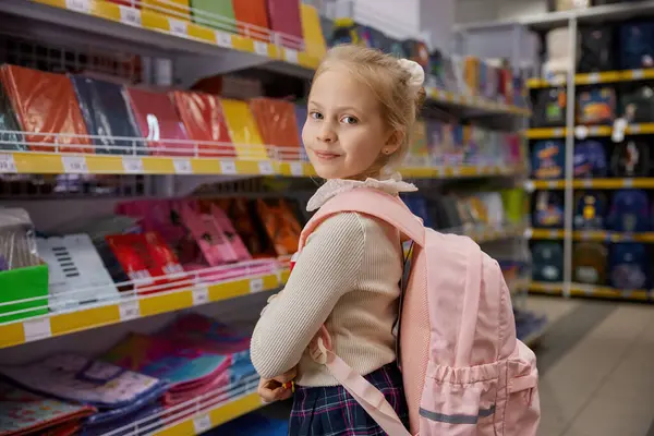 Yeni okul çantasıyla poz veren sevimli küçük kız kırtasiyenin önünde duruyor. Okul malzemelerini süpermarkette seçen bir çocuk.